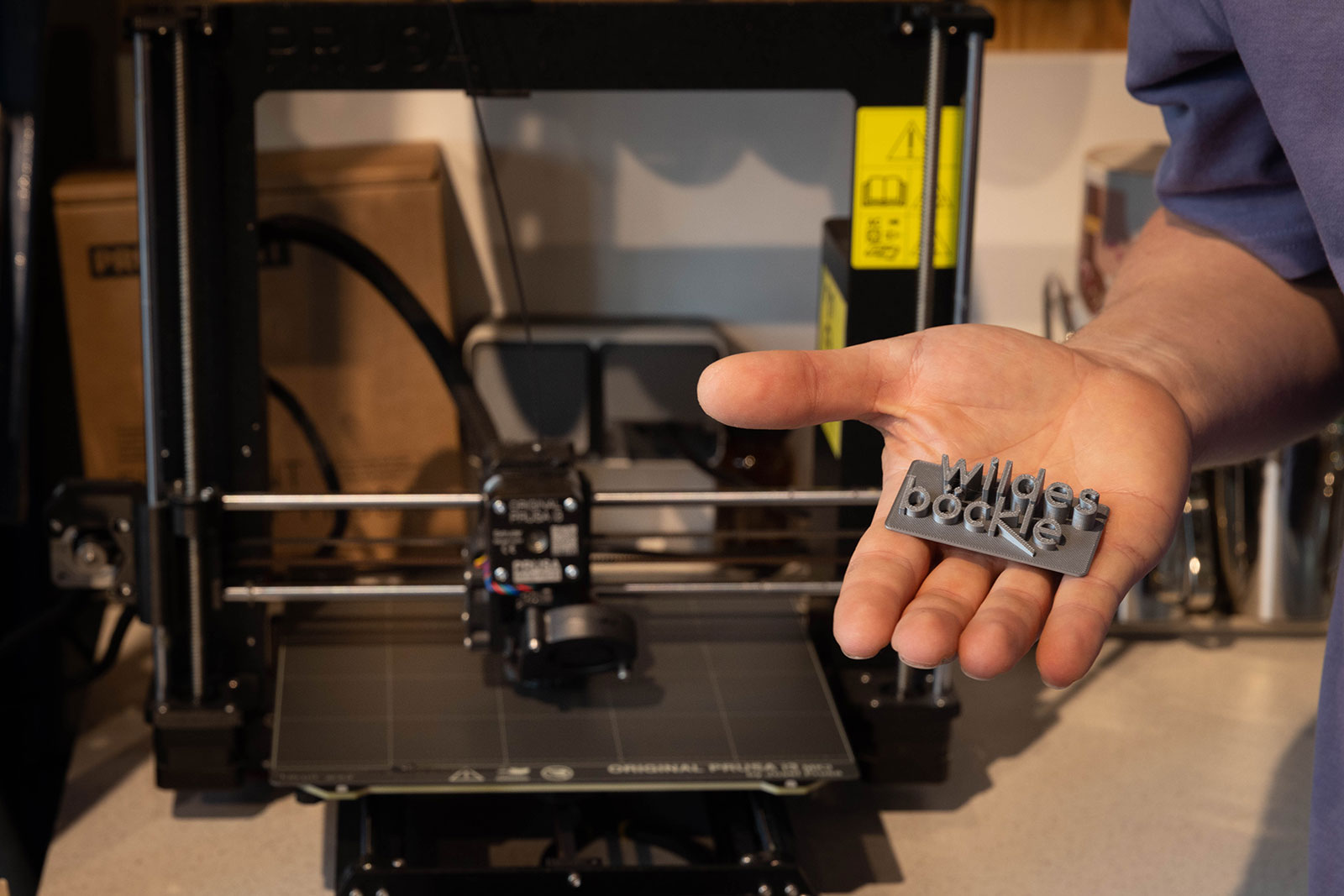 Frank Schwenk zeigt das im 3D-Druck gedruckte wilde böckle-Logo. Im Hintergrund ist der 3D-Drucker zu sehen.